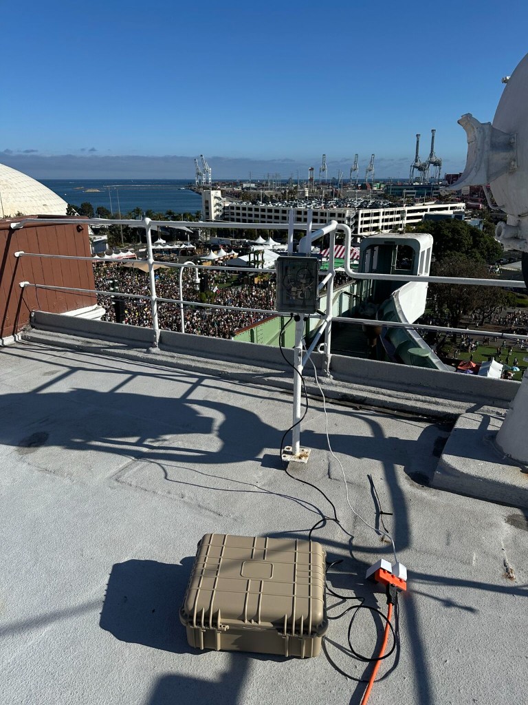 Totem-Air receiver case, antenna, and radio on a rooftop overlooking a festival crowd and harbor
